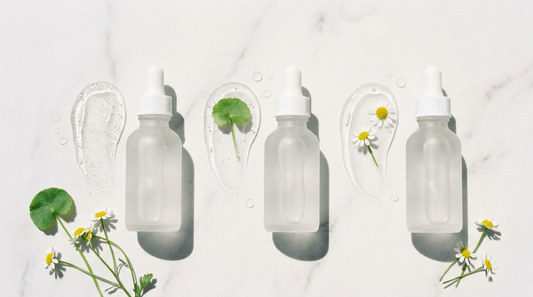 Three frosted dropper bottles with gel textures on white marble with chamomile and centella, representing AHA, BHA and PHA gentle exfoliants for sensitive skin