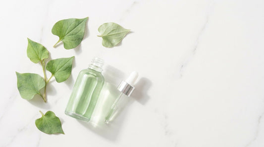 Fresh heartleaf (Houttuynia cordata) leaves and a clear glass dropper bottle with pale green serum on a white marble surface